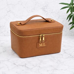 Puede incluir: Un neceser de maquillaje rectangular marrón con cremallera y asa doradas. La funda tiene cierres metálicos dorados y un pequeño parche rectangular con las iniciales doradas "M.L."