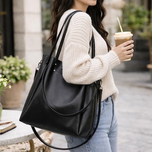 Op de afbeelding: Een zwarte leren tote bag met een schouderriem en een handvat aan de bovenkant. De tas is rechthoekig met een licht trapeziumvormige vorm. De tas wordt over de schouder gedragen.