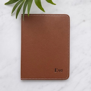 May include: A brown leather passport holder with white stitching around the edges. The name "Eve" is embossed in the lower right corner. A green plant leaf is in the upper left corner.