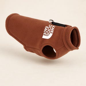 Peut inclure: Un gilet marron pour chien avec le texte "KNE SCIOS FACE" en blanc. Le gilet a un col rond et deux ouvertures pour les pattes avant. Une sangle noire et un anneau métallique sont attachés au sommet du gilet.
