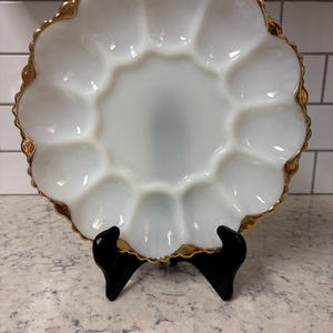 Puede incluir: Un plato para huevos redondo de vidrio lechoso blanco con doce hendiduras circulares dispuestas en anillo alrededor de un centro liso. El plato tiene un borde festoneado con un acabado de borde dorado metálico que enmarca el vidrio.