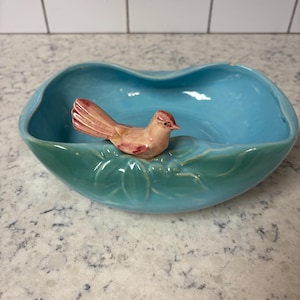 May include: A turquoise ceramic bowl with a bird figurine. The bowl has a leaf-like design around the rim and a light blue interior. The bird is peach-colored with red accents.