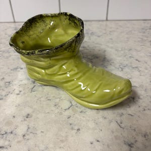 Könnte beinhalten: Ein hellgrüner Keramik-Pflanztopf in Stiefelform mit glänzendem Finish. Der Rand des Stiefels ist schwarz umrandet. Der Stiefel hat ein strukturiertes Design. Er steht auf einer weiß-grauen Oberfläche.