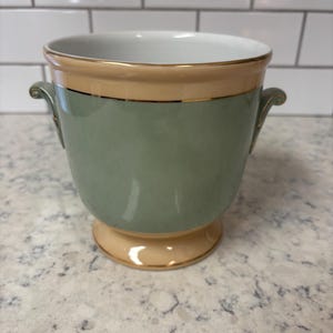 Peut inclure: Un vase en céramique avec un corps vert sauge, une bande beige autour du haut et une base beige. Le vase a deux petites poignées et une bordure dorée. L'intérieur est blanc.
