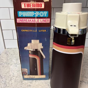 Puede incluir: Un termo Pump-Pot vintage marrón y crema, con una capacidad de 1,2 litros. El termo tiene un diseño retro con una franja roja, naranja y amarilla. Se incluye la caja original, con el nombre del producto y una ilustración del termo.