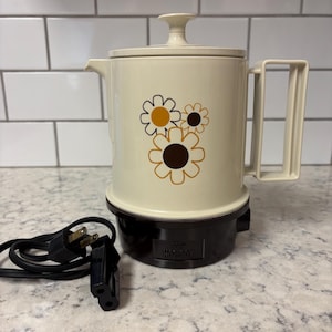 May include: Cream-coloured electric coffee pot with a brown base and handle. The pot features a floral design in brown and yellow. A black power cord and plug are visible. The text "Tom Hotpot" is printed on the base.