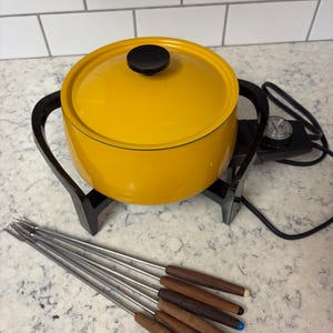 May include: A yellow fondue pot with a black handle and lid knob sits on a black stand. Six fondue forks with wooden handles and coloured tips are in front of the pot. The pot is on a white countertop.