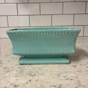 May include: A rectangular, turquoise ceramic planter with a pedestal base. The planter features a wavy, textured design on the sides and a series of small, raised dots along the base. The planter is empty and sits on a speckled countertop.