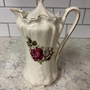 Op de afbeelding: Crèmekleurige keramische creamer met een deksel, met een bloemenontwerp van rode en witte rozen. De creamer heeft een goudkleurige handgreep en tuit, en een decoratieve gouden finial op het deksel. De creamer is ongeveer 15 cm hoog.