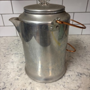 May include: A vintage silver-colored metal coffee pot with a clear glass knob on the lid and a copper-colored handle. The pot has a spout for pouring and is likely used for brewing coffee or tea.