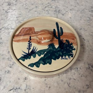 Può includere: Piatto rotondo in ceramica con coperchio raffigurante un paesaggio desertico. Il coperchio ha un pomello ed è dipinto con un cactus, piante del deserto e una mesa nei toni del blu, marrone e beige. La firma dell'artista e l'anno '92 sono visibili.