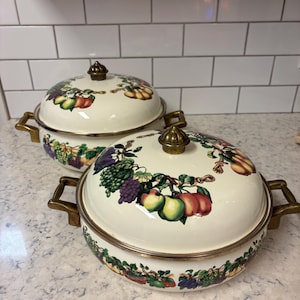 May include: Two off-white enamelware casserole dishes with lids. Each dish features a colorful fruit and vegetable design around the rim and on the lid. The handles and lid knobs are brass-colored.