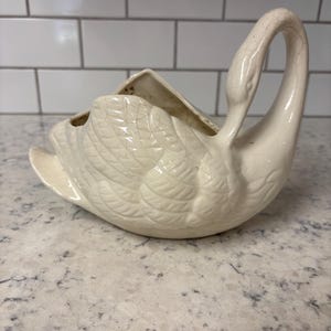 May include: A cream-colored ceramic swan planter featuring a glossy finish with a visible crackle glaze pattern throughout the surface. The swan is depicted with its neck curved upward and wings folded against its body, creating a hollow basin.