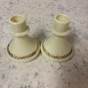 Puede incluir: Dos candelabros color crema con un ribete decorativo dorado alrededor de la base. Los candelabros tienen un diseño similar a un sombrero de copa. Los candelabros son de cerámica o porcelana.