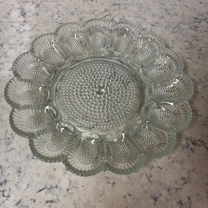 Puede incluir: Plato para servir huevos de vidrio transparente con borde festoneado. El centro del plato tiene un diseño texturizado y punteado. El plato tiene 12 hendiduras en forma de huevo alrededor del borde.