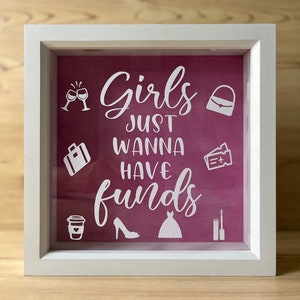 May include: White shadow box with a pink background and the text "Girls just wanna have funds". The text is in white and there are various white icons around the text, including a purse, a suitcase, a pair of high heels, a dress, a glass of wine, a coffee cup, mascara, and an airplane ticket.