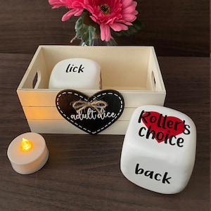 May include: White dice with black text, "lick" and "back" are visible. A black heart with white text, "adult dice" is in the background. A white dice with red heart and black text, "Roller's choice" is in the foreground. The dice are in a wooden crate with a pink flower in the background.