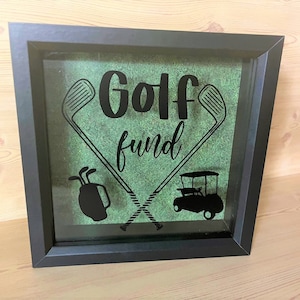 May include: Black framed shadow box with a green background and the text "Golf fund" in black. The box also features a golf cart and golf clubs.