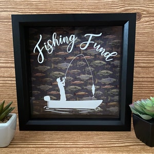 May include: Black shadow box with a white silhouette of a person fishing in a boat. The background is a brown and green pattern of fish. The text "Fishing Fund" is written in white cursive letters.