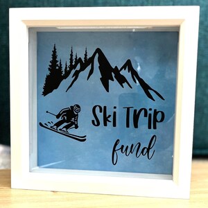 May include: White wooden shadow box with a blue background and a black silhouette of a skier, mountains, and trees. The text "Ski Trip fund" is written in black below the skier.