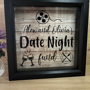 May include: Black shadow box with a woodgrain background. The text "Alex and Olivia's Date Night fund" is written in black. There are icons of a film reel, wine glasses, and a plate with a knife and fork.