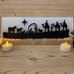 May include: A black silhouette of the Nativity scene on a rectangular glass panel. The scene includes figures of Mary, Joseph, baby Jesus, animals, and the three wise men on camels. The panel is illuminated with a warm glow, and is displayed on a wooden surface.