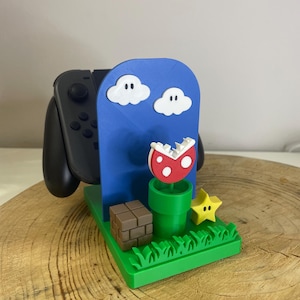 Può includere: Supporto per controller Nintendo Switch stampato in 3D. Presenta uno sfondo blu con nuvole bianche, un fungo rosso e bianco, un tubo verde, una stella gialla e un blocco marrone. Il supporto è su una base verde con un motivo a erba.
