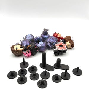 Può includere: Una collezione di piccole figurine colorate e basi circolari nere con pali. Le figurine presentano vari personaggi con tratti del viso e abiti dettagliati. Le basi sono disposte su una superficie bianca.