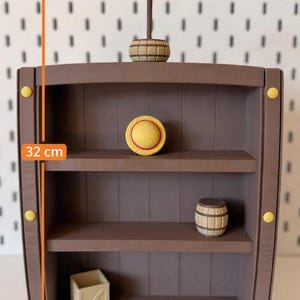 Peut inclure: Une étagère marron à trois niveaux en forme de tonneau mesurant 32 cm de hauteur et 22 cm de largeur. Elle présente des accents circulaires jaunes, un petit chapeau de paille, un tonneau en bois et une caisse claire. Une icône tête de mort est visible.