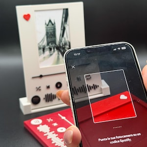 Può includere: Una cornice bianca con un'immagine in bianco e nero di un ponte, un cuore rosso e un codice Spotify rosso. Un telefono sta scansionando il codice. L'ora sul telefono è 11:17.