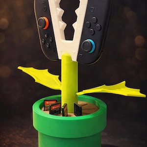 Può includere: Una base a forma di vaso cilindrico verde con uno stelo centrale e due foglie gialle, che sostiene un controller di gioco nero diviso in due metà con strutture bianche simili a denti. La base contiene diverse piccole cartucce di gioco.