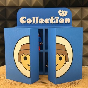 Puede incluir: Un expositor azul con la palabra "Collection" en blanco. El expositor tiene una cara de dibujos animados en la parte delantera y una pequeña estantería en el interior. Las puertas están abiertas, revelando una pequeña figura.