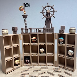 Könnte beinhalten: Ein beigefarbenes Kunststoff-Displayregal mit mehreren Fächern, einem Schiffssteuerrad und einem Mast mit Totenkopfflagge. Kleine Figuren befinden sich in einigen Fächern, beschriftet mit Namen wie Luffy, Vivi und Smoker. Namensschilder liegen davor.