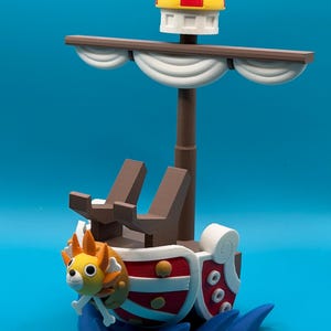 Puede incluir: Un modelo de barco impreso en 3D que presenta un mascarón de proa con forma de león con melena naranja, un casco rojo y blanco, asientos de cubierta marrones, un mástil central con velas blancas, una pequeña cofa amarilla y roja, una bandera negra y olas azules.