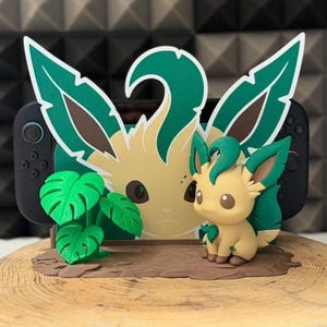 Può includere: Un supporto per console di gioco con una statuetta del personaggio Leafeon color beige e verde, una sagoma piatta abbinata del volto del personaggio e una piccola pianta di monstera verde. Il supporto è progettato per sostenere una console portatile nera.