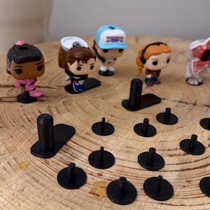 Può includere: Una collezione di piccole statuette di personaggi, tra cui una ragazza in abito rosa, un ragazzo con un cappello bianco e una ragazza con i capelli arancioni. Sono visibili anche basi e perni di plastica nera, il tutto disposto su una superficie di legno.