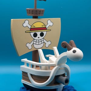 Puede incluir: Un modelo impreso en 3D de un barco con un mascarón de proa de carnero blanco, casco marrón y una vela color canela que muestra una calavera con sombrero de paja amarillo y banda roja, sobre una base azul que representa olas del mar.
