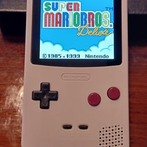 May include: A white handheld gaming console with a backlit screen displaying the Super Mario Bros. Deluxe title screen. The device features a black directional pad, two dark pink A and B buttons, and Select and Start buttons below the screen.