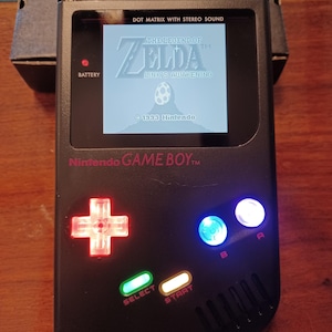 May include: A black handheld video game console with a backlit screen displaying The Legend of Zelda: Link's Awakening. The device features a glowing red directional pad, glowing blue B button, glowing white A button, and illuminated green and yellow buttons.