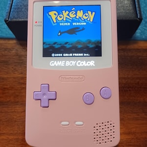 May include: A pink Game Boy Color handheld gaming console featuring a screen displaying the Pokémon Silver Version title screen with the text 2000 GAME FREAK inc. The device has a purple directional pad, purple A and B buttons, and white Select and Start buttons.