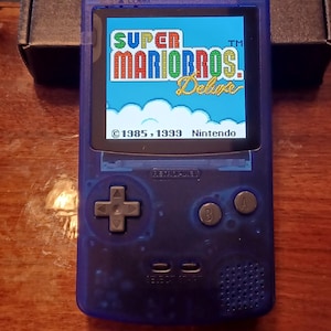 May include: A translucent blue handheld video game console featuring a backlit screen displaying the Super Mario Bros. Deluxe title screen with the text 1985, 1999 Nintendo. The device includes a directional pad, A and B buttons, and start buttons.