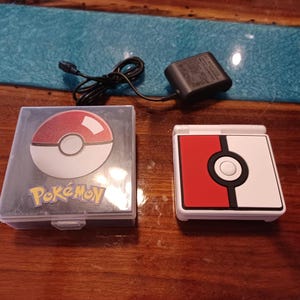 May include: A handheld gaming console featuring a red and white Pokeball design, accompanied by a matching clear plastic storage case with a Pokeball graphic and the word Pokemon, along with a black power adapter with a thin black charging cable.
