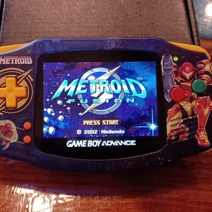 May include: A blue and yellow Game Boy Advance handheld console featuring a Metroid Fusion theme. The screen displays the Metroid Fusion title screen with the text Press Start and 2002 Nintendo. The console has a yellow D-pad and buttons.