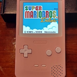 May include: A light pink handheld gaming console with a backlit screen displaying the Super Mario Bros. Deluxe title screen. The device features a directional pad, A and B buttons, Select and Start buttons, and a speaker grille. The text RETROPIXEL is embossed above the directional pad.