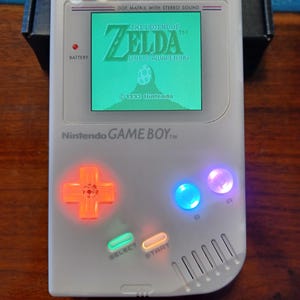 May include: A white Nintendo Game Boy handheld console with a lit screen displaying "The Legend of Zelda: Link's Awakening." The directional pad glows orange, and the A and B buttons glow blue and purple. The console has the text "Nintendo GAME BOY" on the bottom.