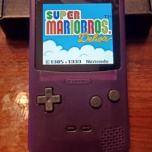 May include: A purple handheld video game console featuring a backlit screen displaying the Super Mario Bros. Deluxe title screen. The device includes a directional pad, A and B buttons, select and start buttons, and a speaker grille on the front.