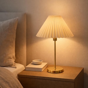May include: A bedside table lamp with a pleated cream-colored shade and a gold-colored base and stem. The lamp is on a wooden nightstand next to a bed with a beige comforter and pillow. A small white bowl and a book are on the nightstand.