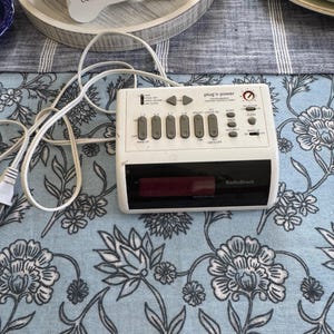 May include: A white RadioShack plug n' power clock radio with a digital display and multiple buttons. The clock is on a floral patterned tablecloth. A decorative dog bone with the words "live laugh bark" is in the background.