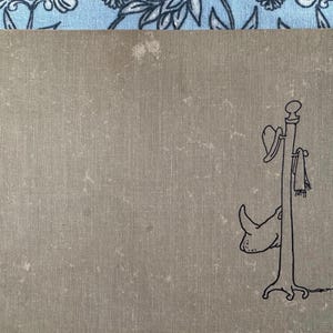 May include: A hardcover book with a tan cover. A black line drawing of a rhino head and a coat rack with a hat and scarf is on the right side of the book. The book is on a blue floral patterned background.