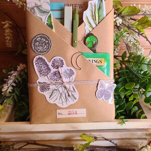 Puede incluir: Un paquete de papel marrón atado con hilo, adornado con pegatinas botánicas, un bolígrafo verde y un amuleto de trébol de cuatro hojas. El paquete contiene artículos de papelería y una bolsita de té. El número 251 está impreso en una etiqueta.